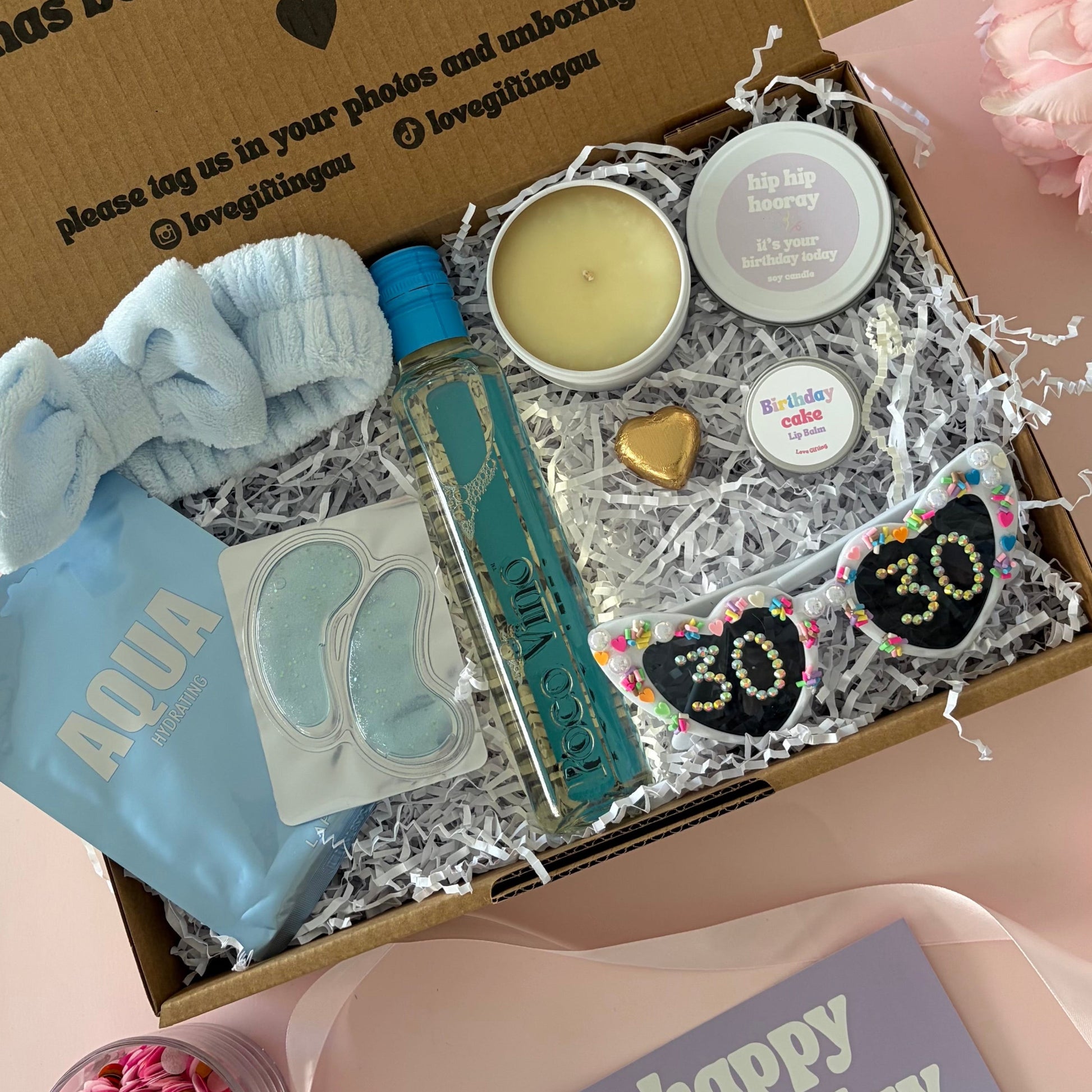 A curated 30th birthday gift box . The box contains a light blue baby headband a mini  bottle of sav blanc, a small candle, lapcos face mask , under eye mask ,lipbalm and age '30'  sunglasses 