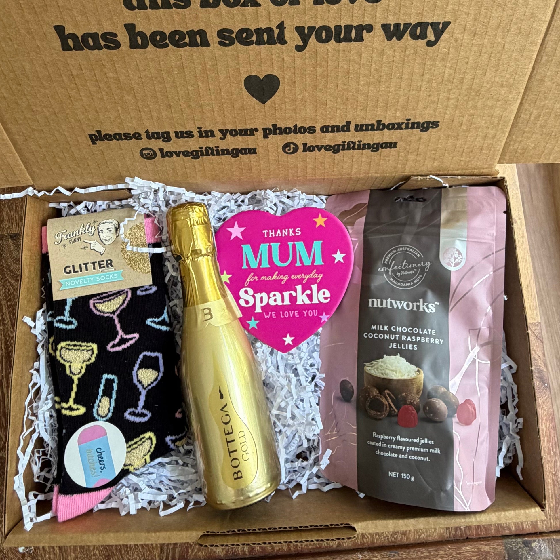 Gift box with various items including a bottle, card, and snacks on a wooden surface.
