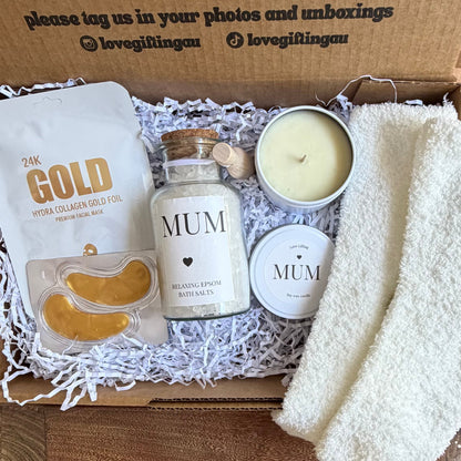 Gift box with products including a bottle labeled 'MUM', a candle, and a towel on a wooden surface.