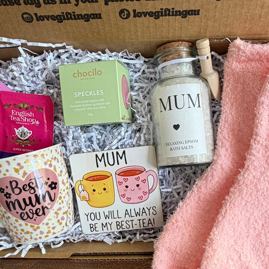 Gift box with 'MUM' bottle, pink socks, and other items on a wooden surface.