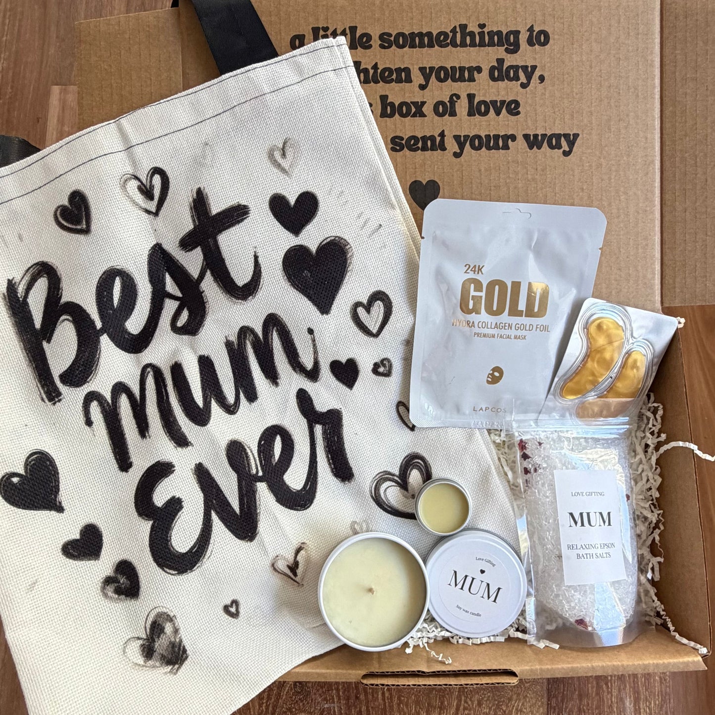 Gift set with 'Best Mum Ever' bag, gold packets, and skincare items on a wooden surface.