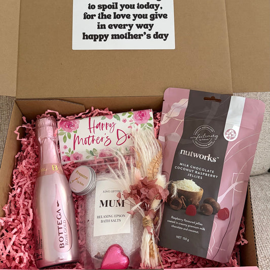 Gift box with Mother's Day items including a bottle, card, and small plant on a beige surface.