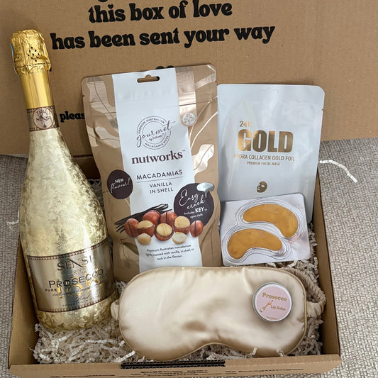 Gift box with champagne, macadamia nuts, and eye masks on a textured surface.