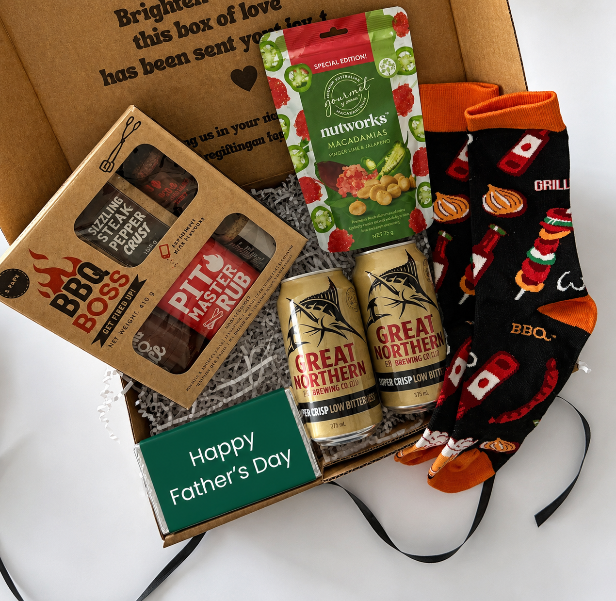 A curated Father's Day gift box filled with BBQ-themed items: a boxed BBQ rub and sauce, a bag of Mt. Vernon fruit snacks, two cans of Great Northern beer, and a pair of black socks with orange cuffs and footprints, arranged in a brown cardboard box with a 'Happy Father's Day' card.