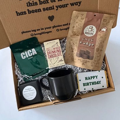 A curated gift box set shown from above inside an open brown cardboard box with printed message on the inside lid. The set includes a black ceramic mug, a green bag of CICA coffee, a chocolate bar, a small boxed item, and a birthday card, arranged in white shredded paper.