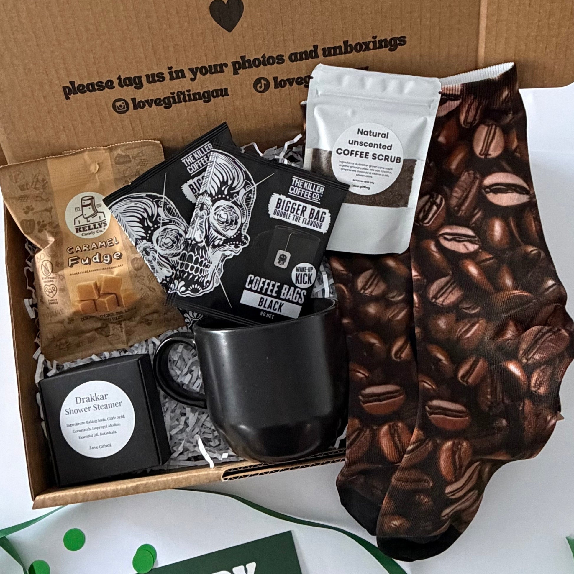 A curated coffee gift box set displayed on a white surface, featuring a brown cardboard box printed with a message, a black ceramic mug, a black coffee mug sleeve with a coffee bean pattern, coffee bags, a green 'happy birthday' card, and green ribbon confetti.