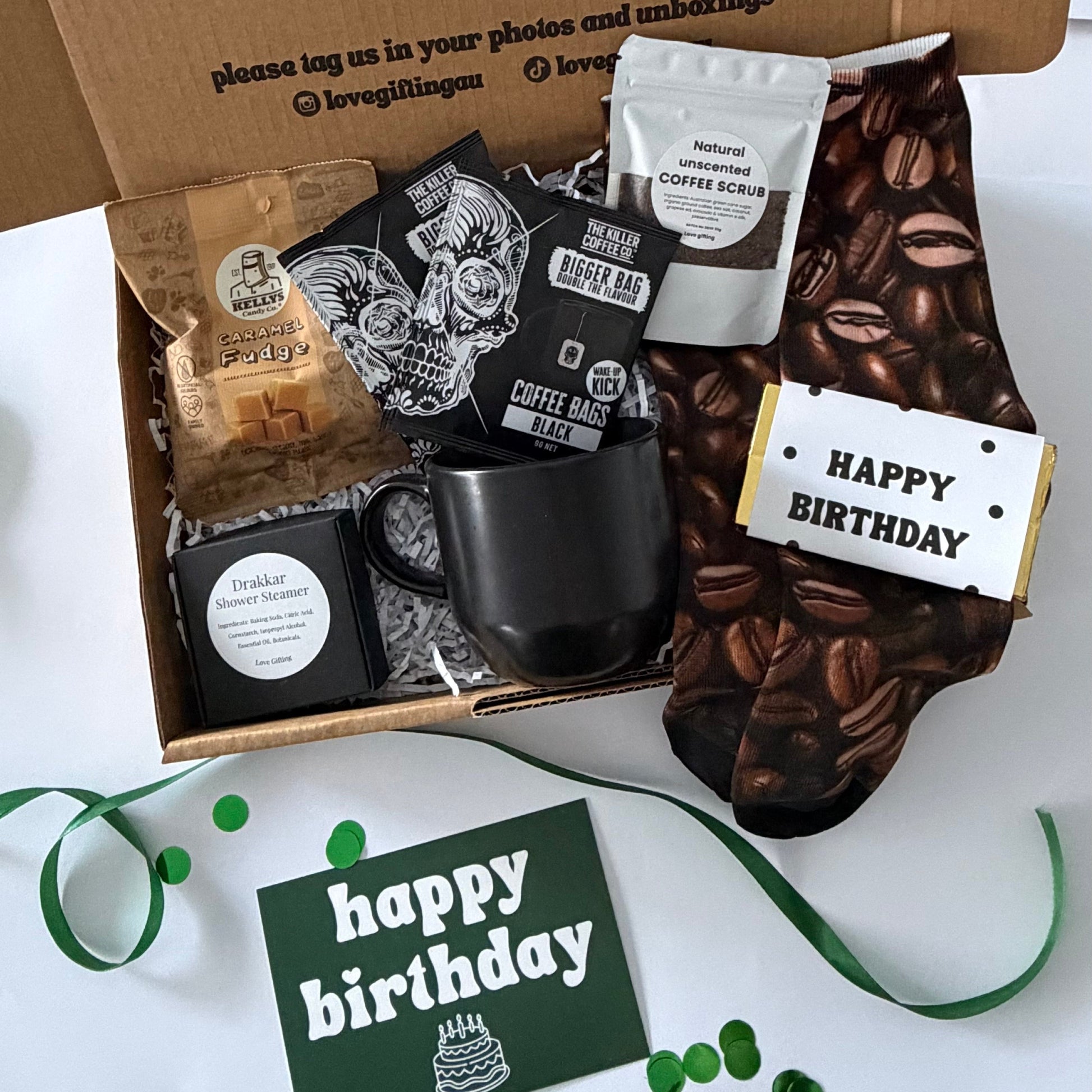 A curated coffee gift box set displayed open on a white surface. The brown cardboard box lid has printed text, and the box contains a black coffee mug, packaged coffee beans, a bag of coffee, a coffee bean patterned sock, and a small card that says Happy Birthday.