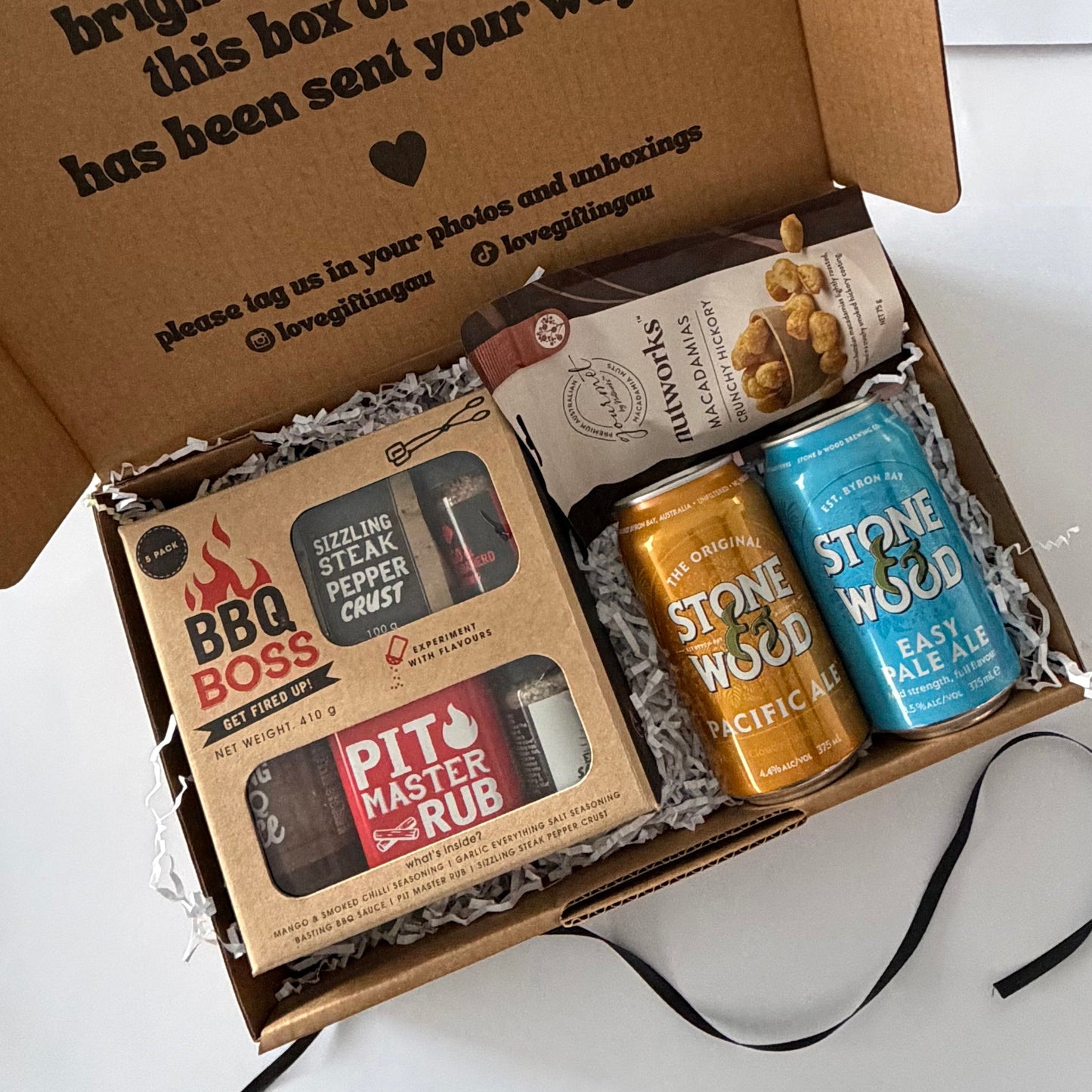 An open cardboard gift box filled with packaged items on a white surface. Visible contents include a boxed BBQ Boss steak rub, a bag of pecans, and two Stone Wood canned ales in orange and blue.