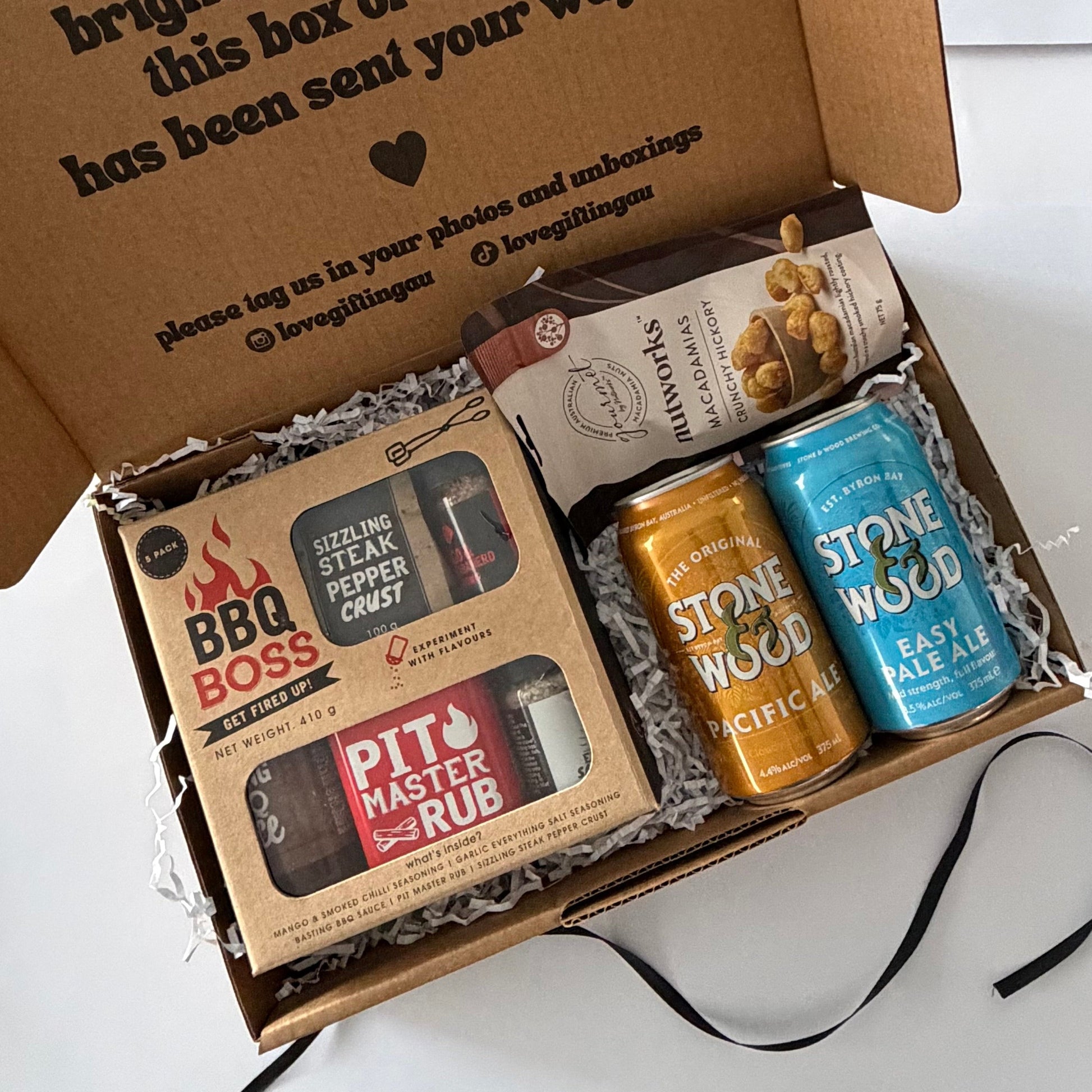 An open cardboard gift box filled with packaged items on a white surface. Visible contents include a boxed BBQ Boss steak rub, a bag of pecans, and two Stone Wood canned ales in orange and blue.