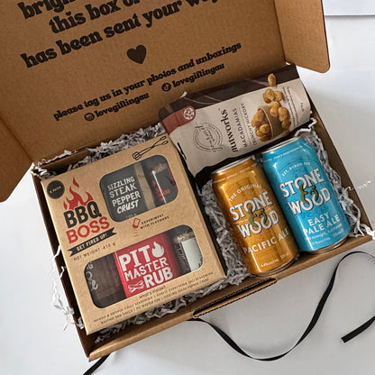 An open cardboard gift box filled with packaged items on a white surface. Visible contents include a boxed BBQ Boss steak rub, a bag of pecans, and two Stone Wood canned ales in orange and blue.