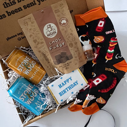 A curated gift box filled with treats and apparel on a white background. The box contains a chocolate bar, a bag of chocolate fudge, a blue 'Happy Birthday' card, and a pair of black BBQ-themed socks with orange accents.