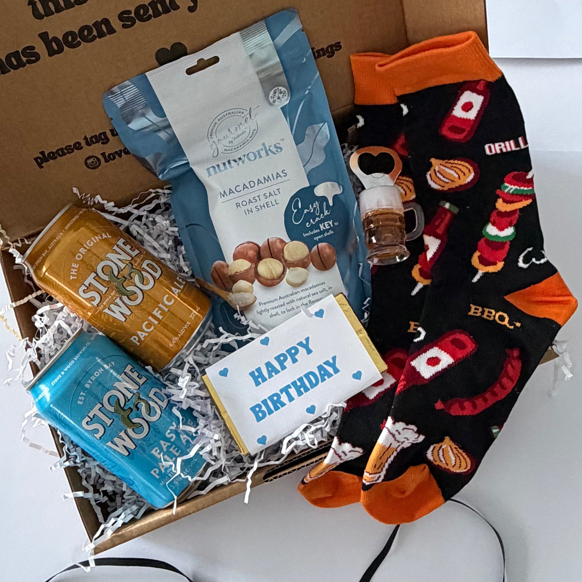 A curated gift box set displayed in an open brown cardboard box on a white surface. The set includes a blue bag of gourmet macarons, a gold-wrapped chocolate, a blue wrapped item, a small bottle, a 'Happy Birthday' card, and a pair of black crew socks with orange cuffs and red/orange holiday-themed graphics.
