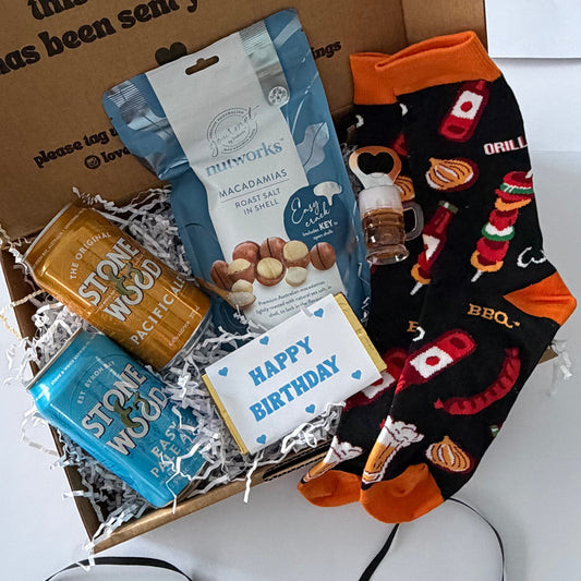 A curated gift box set displayed in an open brown cardboard box on a white surface. The set includes a blue bag of gourmet macarons, a gold-wrapped chocolate, a blue wrapped item, a small bottle, a 'Happy Birthday' card, and a pair of black crew socks with orange cuffs and red/orange holiday-themed graphics.
