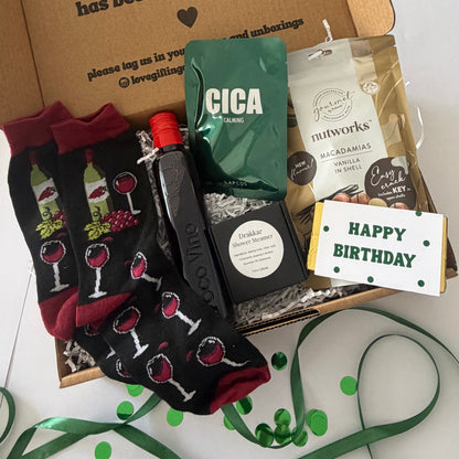A curated gift box set containing a pair of black crew socks with a wine glass pattern, a bottle of olive oil, a green bag of Cica flour, a bag of Nutworks Macadamia Flavouring, and a 'Happy Birthday' chocolate bar, all arranged in a cardboard box with green ribbon and confetti.
