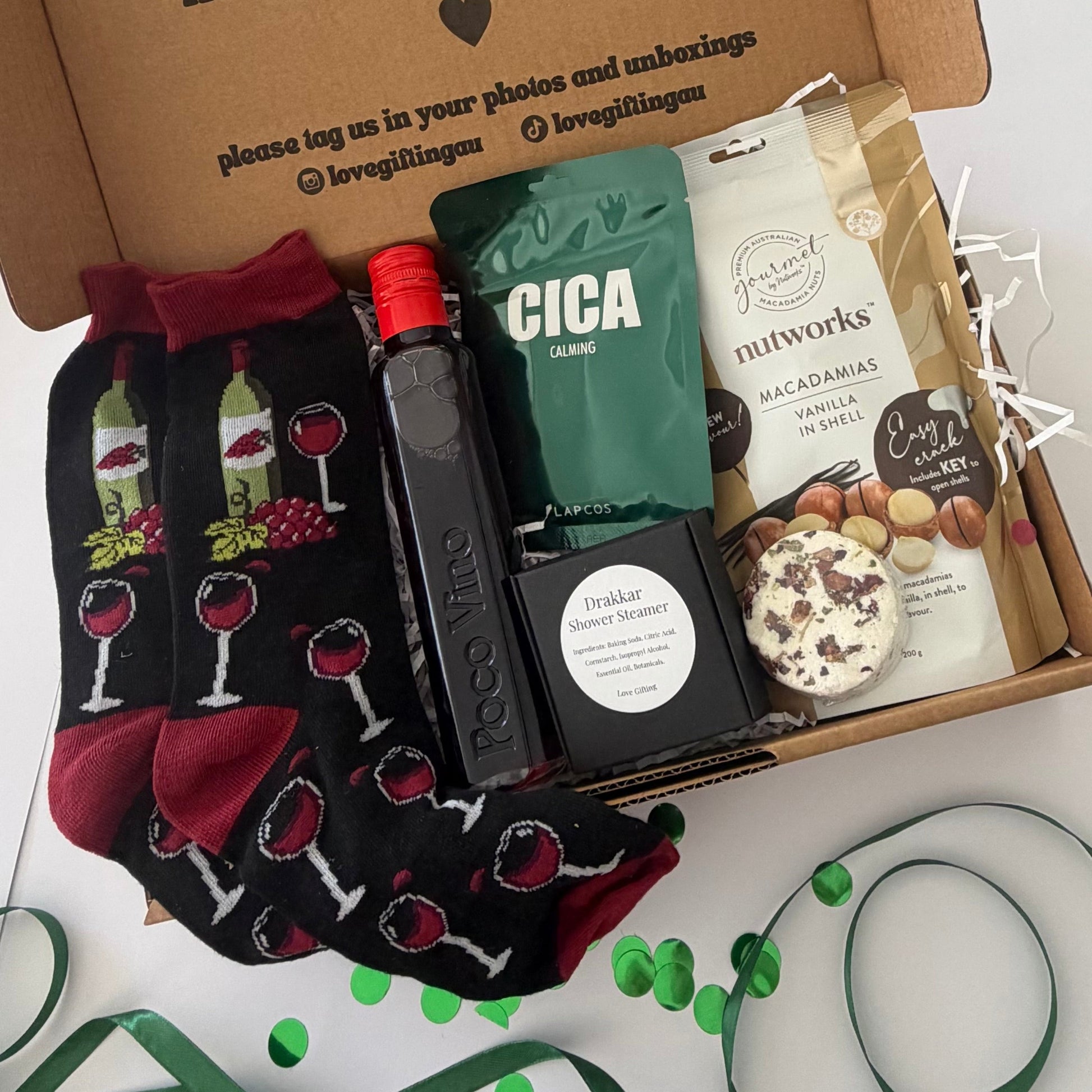 A curated gift box set displayed inside an open brown cardboard box on a white surface. The set includes black crew socks with a red wine glass and grape pattern, a bottle of Cica coffee, a black box of chocolate-covered nuts, a bag of Nutworks macadamia nuts, and a bag of chocolate chip cookies.