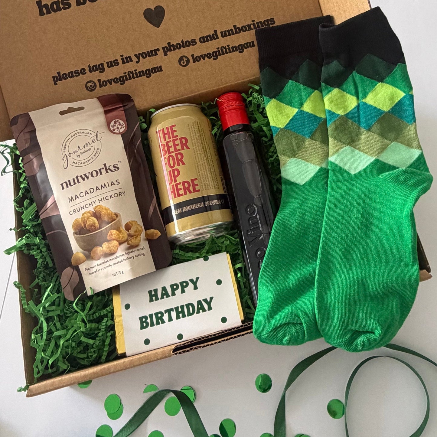 A curated gift box set on a white surface, featuring a pair of green argyle socks, a bag of Nutworks Macadamia snacks, a can of The Beer Brand IPA, a black bottle, and a 'Happy Birthday' card nestled in green paper filler inside an open cardboard box.
