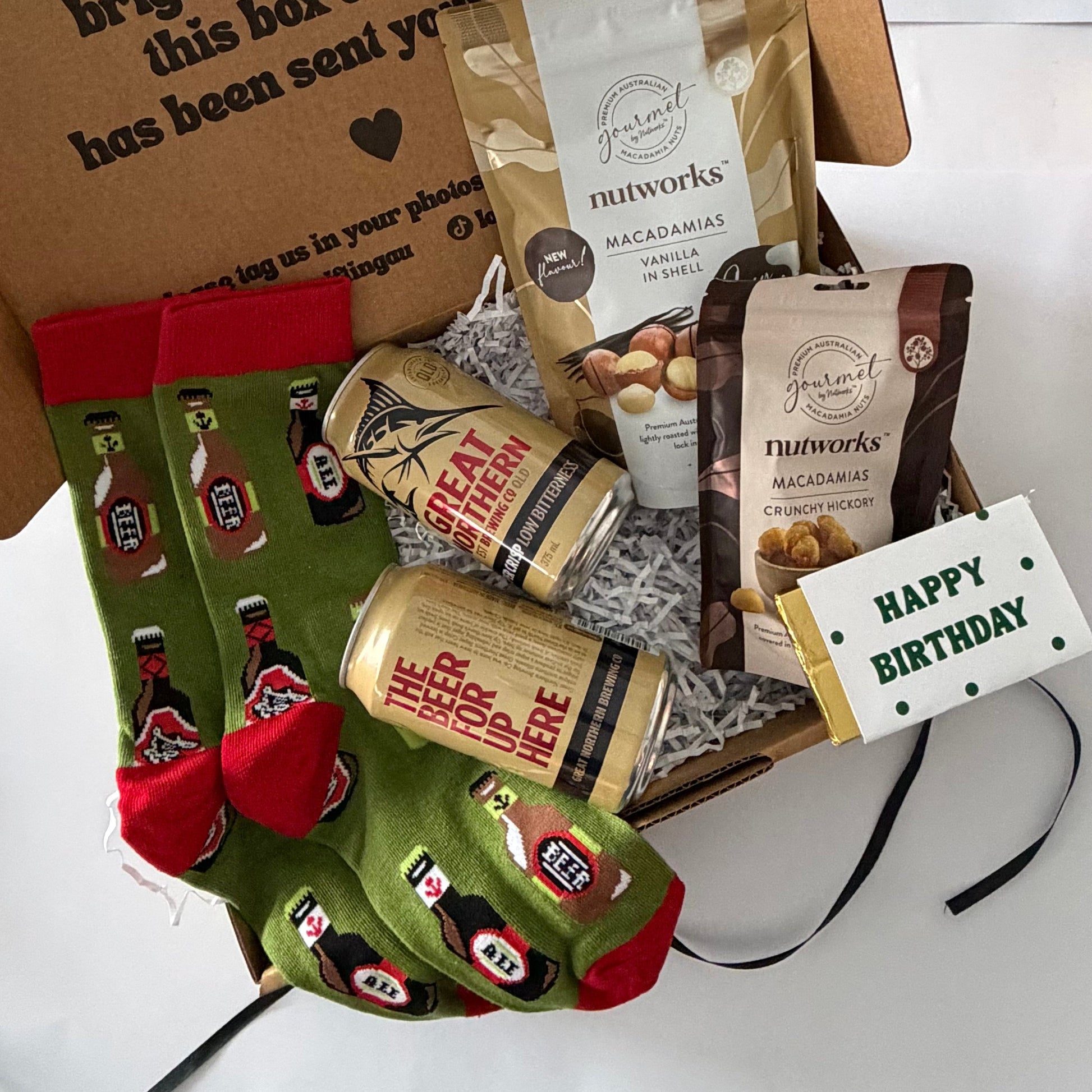 A curated gift box filled with snacks and novelty socks. The box contains Nutworks macadamia and chocolate chip bags, a can of Miller High Life, a 'Happy Birthday' card, and a pair of green crew socks with red heels and toes featuring a red and black beer bottle pattern.
