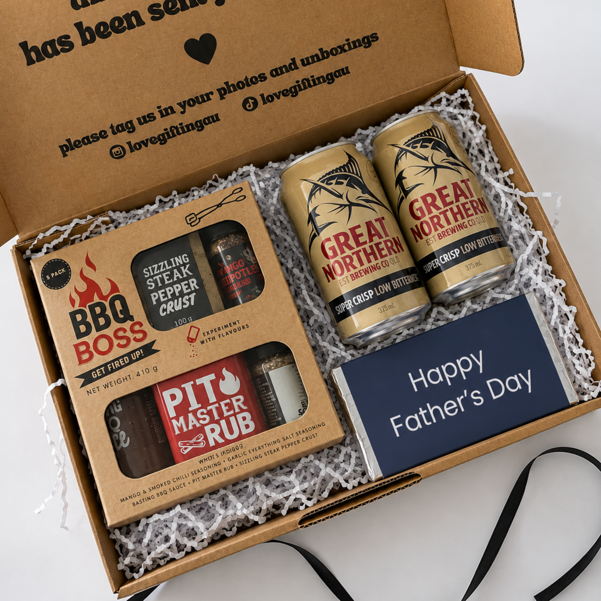A curated Father's Day gift box from Lovegiggins, featuring a cardboard interior printed with a message, two cans of Great Northern beer, a BBQ Boss rub, a Pit Master Rub, and a blue Happy Father's Day card nestled in white shredded paper.