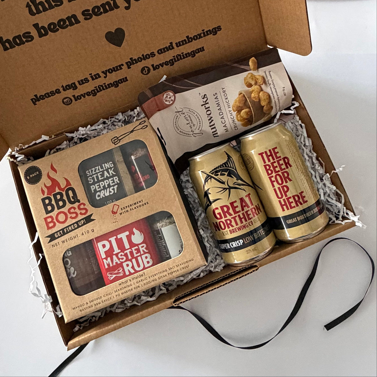 A curated gift box set containing a 'BBQ BOSS' seasoning rub kit, a bag of 'STRAWBERRY' blueberries, and two gold 'GREAT NORTHERN' beer cans, arranged in a brown cardboard box with shredded paper filler.