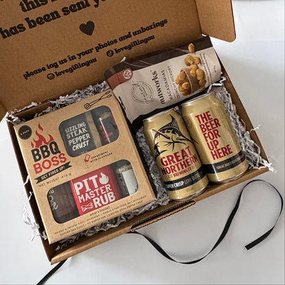 A curated gift box set containing a 'BBQ BOSS' seasoning rub kit, a bag of 'STRAWBERRY' blueberries, and two gold 'GREAT NORTHERN' beer cans, arranged in a brown cardboard box with shredded paper filler.