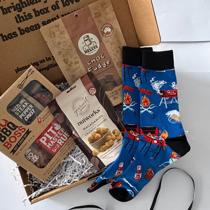 A curated gift box set on a white background, featuring a brown cardboard box with printed text, a bag of chocolate fudge, a bag of Pita Bites, and a pair of blue crew socks with a campfire and grill pattern.