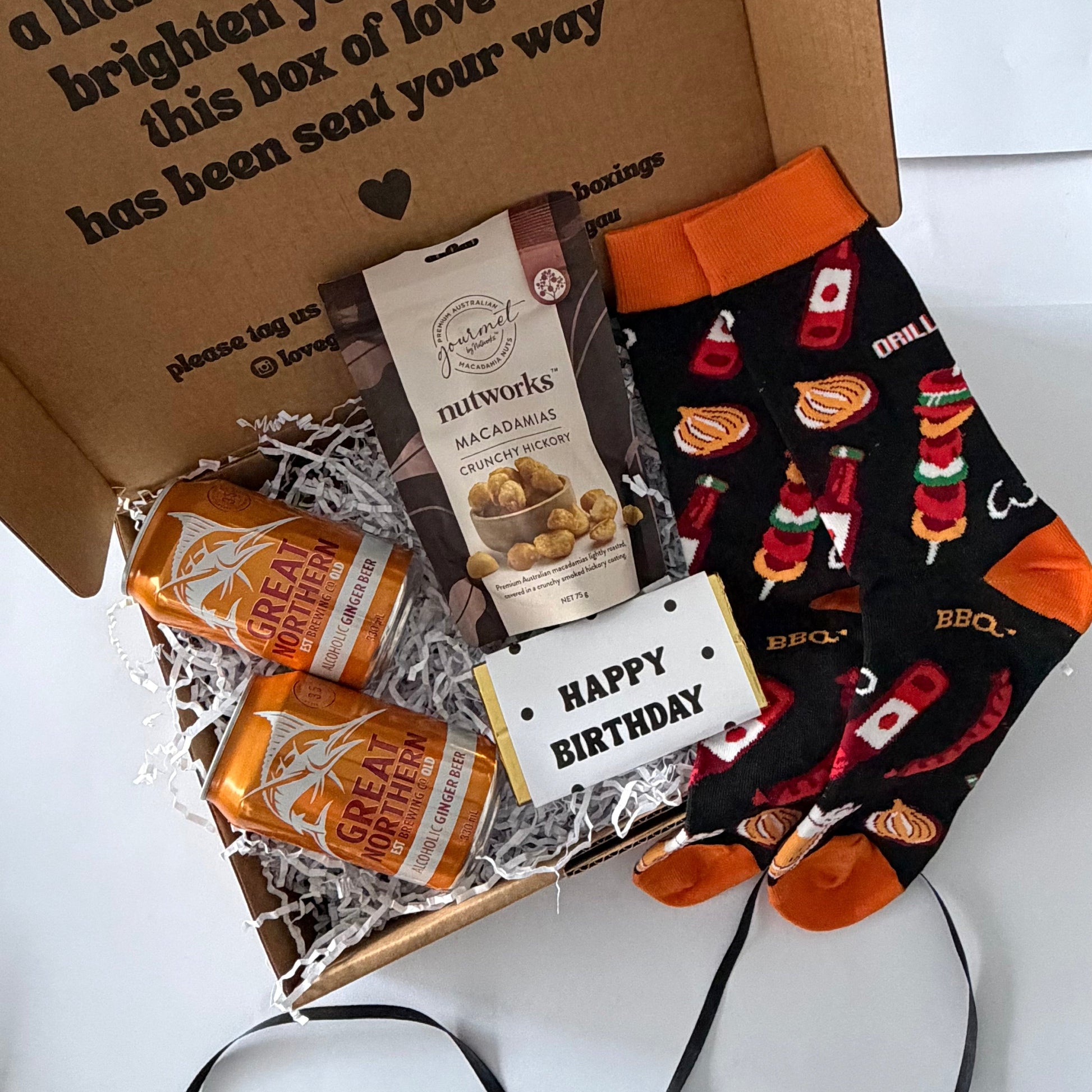 A curated gift box set on a white background, featuring a brown cardboard box with a printed message, two bags of Nutworks Macadamia snacks, two orange-and-white striped soda cans, and a pair of black socks with a red and orange BBQ grill pattern and orange cuffs.