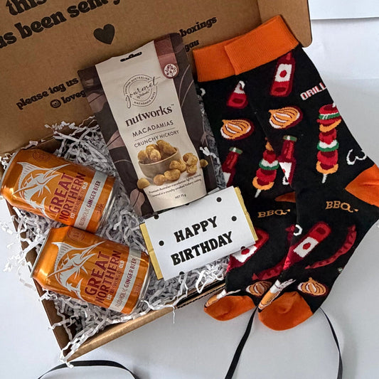 A curated gift box set on a white background, featuring a brown cardboard box with a printed message, two bags of Nutworks Macadamia snacks, two orange-and-white striped soda cans, and a pair of black socks with a red and orange BBQ grill pattern and orange cuffs.