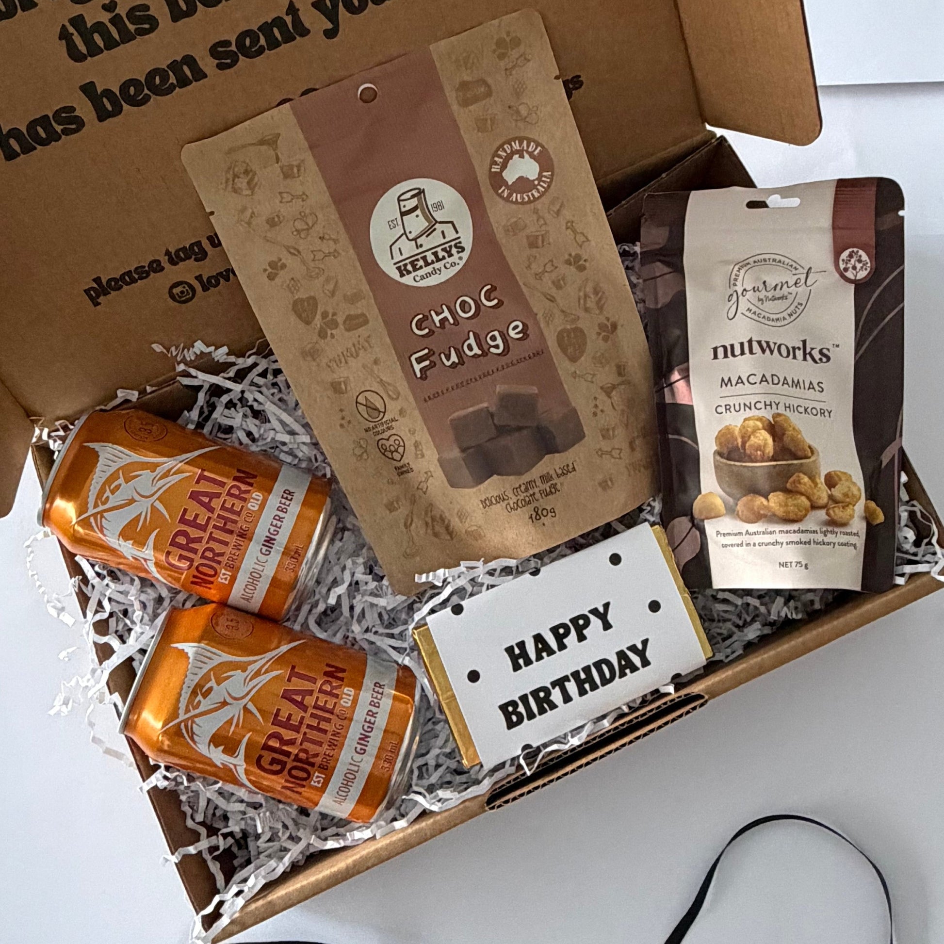 A curated gift box set on a white surface, featuring a brown cardboard box printed with a birthday message, filled with shredded paper and snacks: a bag of Choc Fudge, a bag of Nutworks Macarons, two orange cans of Great Britain, and a small white card that says Happy Birthday. A black ribbon is draped in front of the box.
