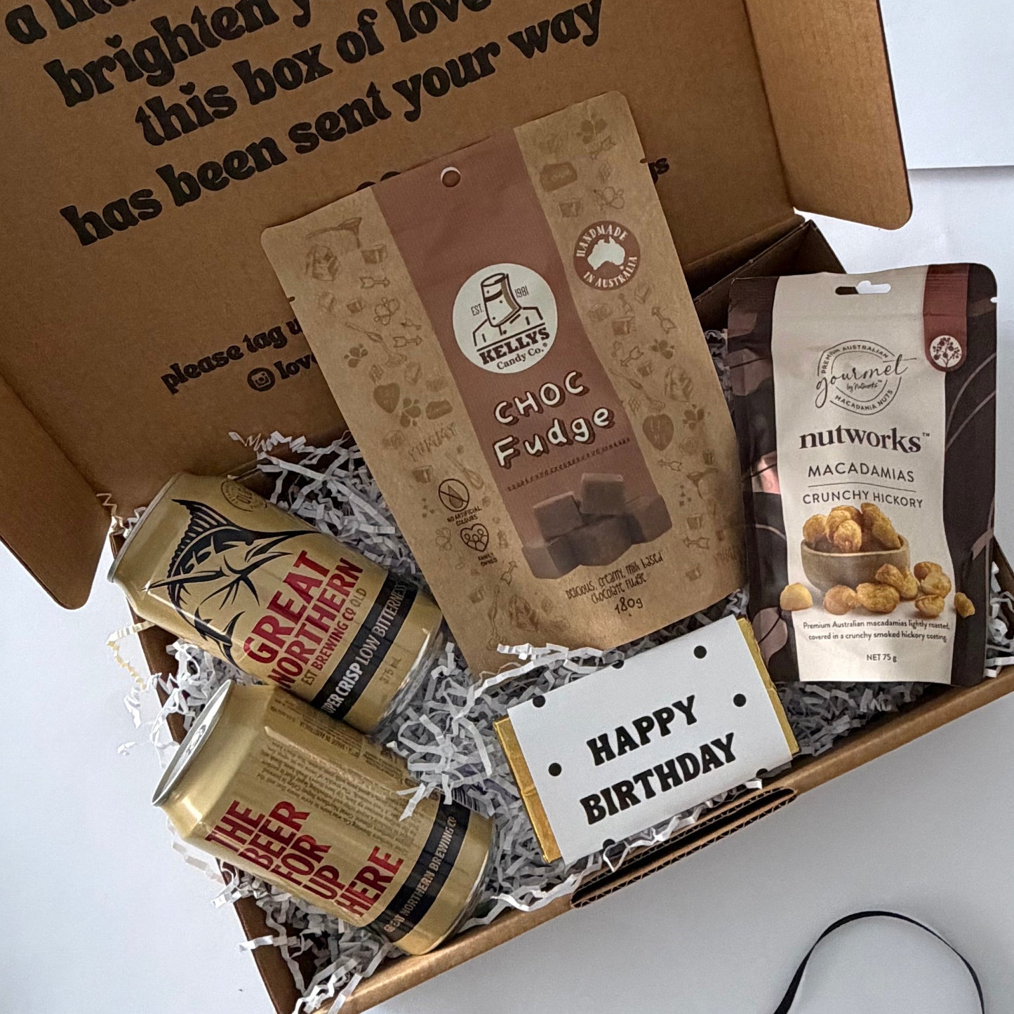 A curated birthday gift box filled with treats and a handwritten-style card. The open brown cardboard box has printed text on the inside lid and is arranged on a white surface with shredded paper filler.