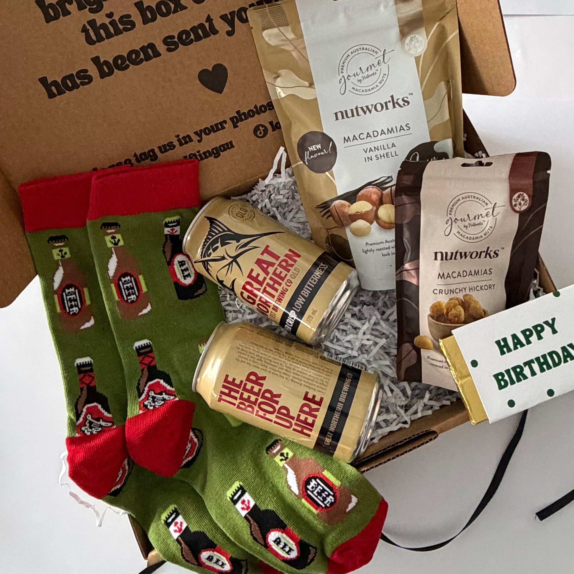 A curated gift box filled with snacks and novelty socks. The box contains Nutworks macadamia and chocolate chip bags, a can of great northern beer a 'Happy Birthday' cahocolate bar and a pair of green crew socks with red heels and toes featuring a red and black beer bottle pattern.