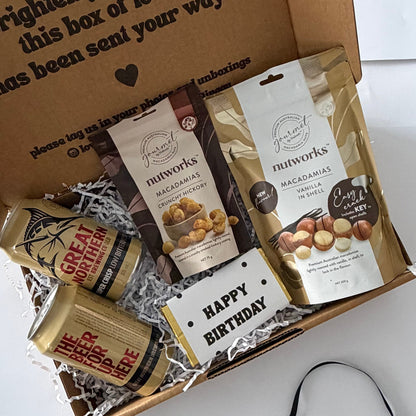 A curated gift box set on a white background, featuring a brown cardboard box with printed text, assorted packaged snacks including mineworks chocolate and Great British Treats chocolate, a small white card reading 'HAPPY BIRTHDAY', and a black ribbon.