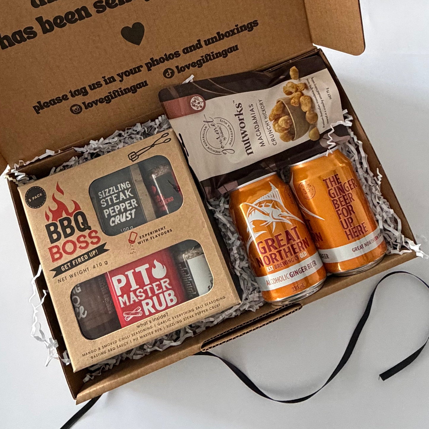 An open cardboard gift box filled with curated items on a white surface. Visible contents include a BBQ Boss rub and pepper spray set, a bag of marshmallows, and two orange canned drinks.