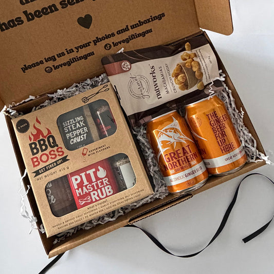 An open cardboard gift box filled with curated items on a white surface. Visible contents include a BBQ Boss rub and pepper spray set, a bag of marshmallows, and two orange canned drinks.