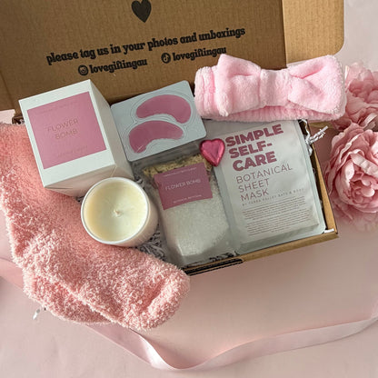 A gift box set containing a pink eye mask sheet pack, a pink bath bomb, a pink candle, a pink towel, and a pink satin bow, arranged on a pink gift bag next to a brown kraft card that reads 'a little something to brighten your day.