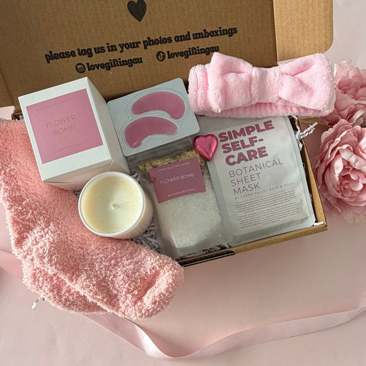A gift box set containing a pink eye mask sheet pack, a pink bath bomb, a pink candle, a pink towel, and a pink satin bow, arranged on a pink gift bag next to a brown kraft card that reads 'a little something to brighten your day.