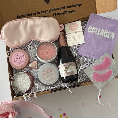 A curated beauty gift box  filled with spa items: a pale pink satin eye mask, pink under eye mask, lapcos face mask sheet, epsom bath salt tin, chocolate heart, Yellow Tail Sparkling Rosé  and happy birthday bracelet