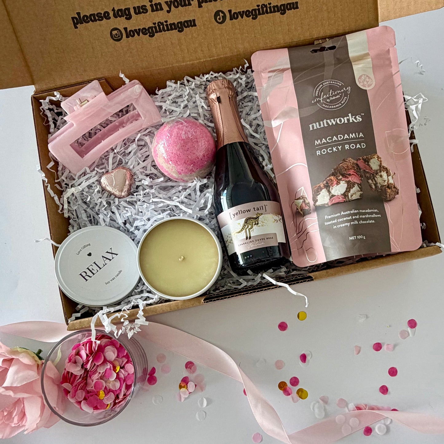 A curated pamper  gift box  includes a starwberry bath bomb, a relax candle, a pink chocolate heart,  a small bottle of sparkling rosé, , a bag of Nutworks macadamia rocky road, and a pink claw hair clip