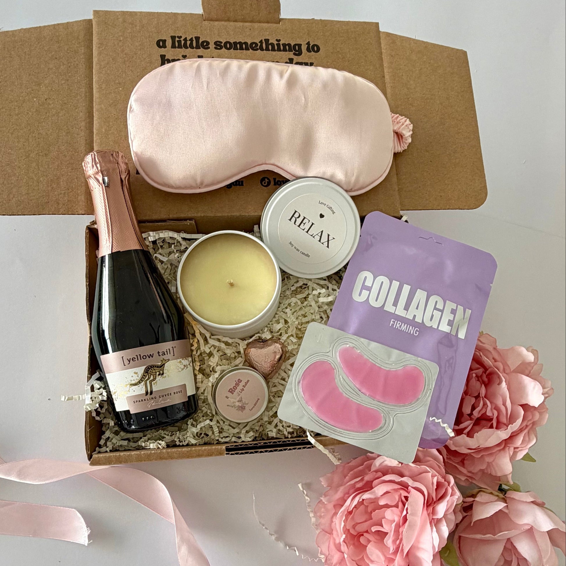 A curated gift box set on a white surface, featuring a light pink satin sleep mask, a bottle of Prosecco, a candle,  lapcos collagen facemask sheet, prosecco lipbalm, chocolate heart