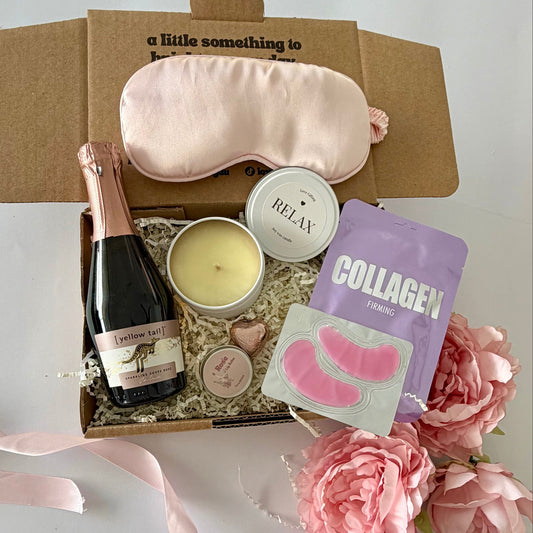 A curated gift box set on a white surface, featuring a light pink satin sleep mask, a bottle of Prosecco, a candle,  lapcos collagen facemask sheet, prosecco lipbalm, chocolate heart