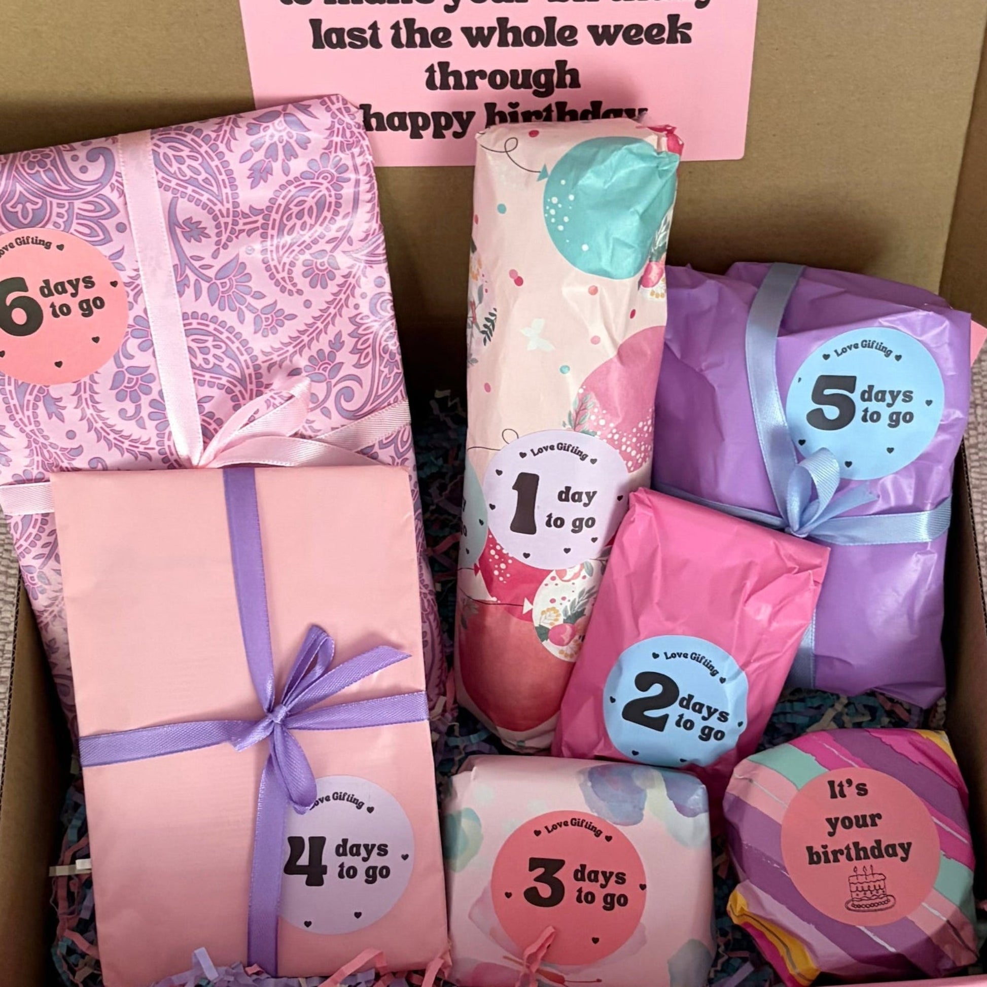 Box of birthday-themed products with pink and purple packaging on a beige surface.