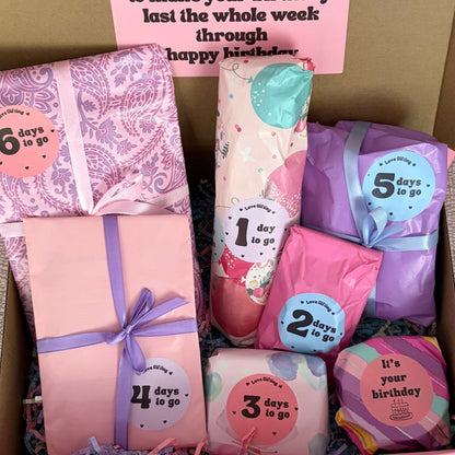 Box of birthday-themed products with pink and purple packaging on a beige surface.