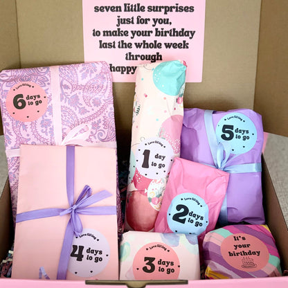 Box of wrapped gifts with numbered stickers, pink box with purple ribbon, and a card on a neutral background