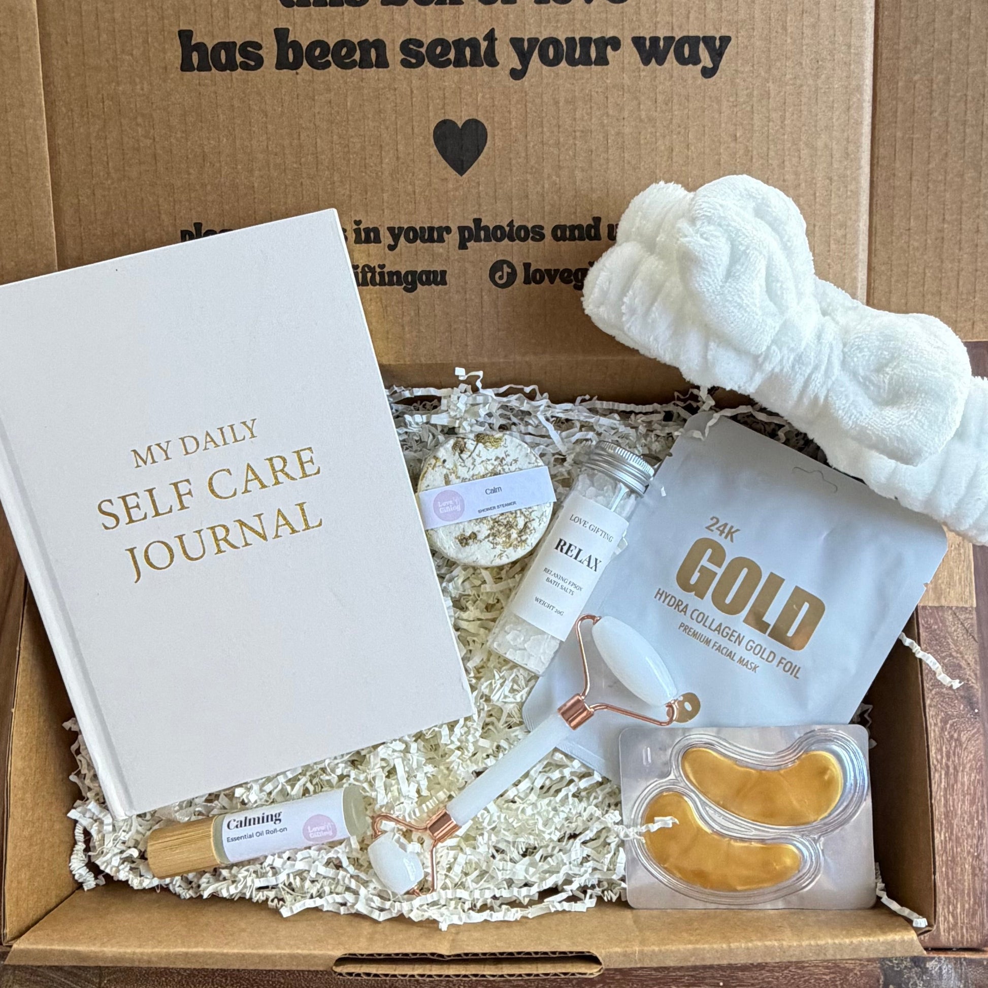 Box of self-care items including a journal, eye masks, and a bottle labeled 'Gold' on a wooden surface.