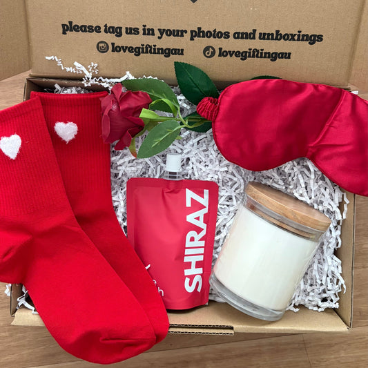 Box of valentines gifts made with love with red socks, a candle, and a bottle of Shiraz wine on a wooden surface.