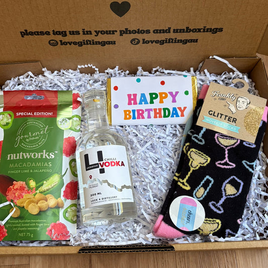 Gift box with birthday items including a bottle of vodka, snacks, and socks on a wooden surface.
